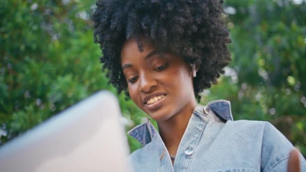 黑头发的女人坐在绿树下的笔记本电脑旁进行视频拍摄 美丽微笑的非洲裔美国女孩在户外对着电脑摄像头说话的肖像 可爱的自由职业者在网上发言 — 图库视频影像