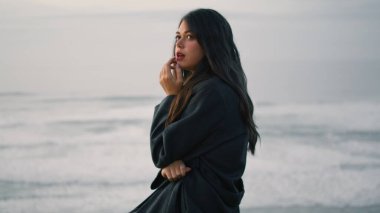 Silhouette of tranquil young woman enjoying seascape at night close up. Attractive dreamy model posing in front gray foamy ocean waves at twilight. Long-haired fashion girl sitting beach at dusk.