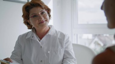 Orta yaşlı, gözlüklü kadın doktor estetik cerrahi cilt bakımı için hastaya danışmanlık yapıyor. Tıp kürklü profesyonel hekim masaya oturup tedaviyi yakından tartışıyor..