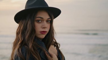 Charming young woman with dark magnetic eyes standing coast overcast autumn day close up. Portrait of long-haired attractive brunette looking camera in front gray ocean. Calm nature beauty concept.