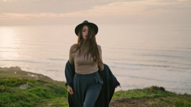 Stylish sensual model walking on green hill near beautiful calm ocean close up. Attractive confident woman posing at amazing sunset autumn evening. Fashionable long-haired girl putting on dark coat.