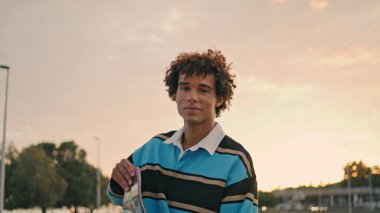 Stylish student holding skate at evening city portrait. Young man looking camera alone at sunset background. Smiling skateboarder posing at urban place outdoor. Contemporary modern lifestyle concept