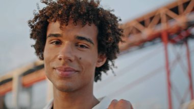 Smiling teenager posing sunset street portrait. Curly hair model staring at urban bridge background. Positive guy looking camera at cloudy evening closeup. Calm hipster face relaxing alone outdoors