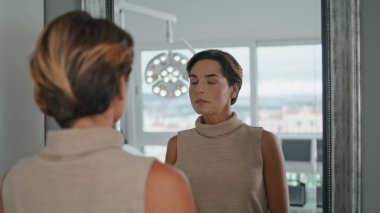Orta yaşlı bir kadın modern kozmetoloji kliniğinde aynaya bakıyor. Çekici zarif bayan güzellik salonunda yaşlanmayı önleyici estetik prosedürler uyguluyor. Yüz derisini inceleyen istemci.