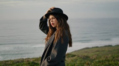 Attractive young model posing holding hat at ocean coast gloomy autumn evening close up. Serious confident woman wearing elegant coat standing alone on green grass seashore. Brunette looking scenery.
