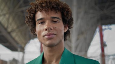 Elegant guy smiling camera at street closeup. Positive young man walking at summer day. Curly hair model posing at urban settings portrait. Handsome teen face looking boldly outdoors. People concept