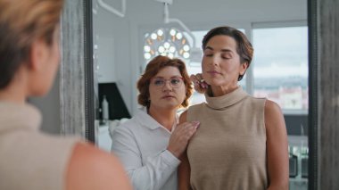 Kendine güvenen kozmetolog müşterisi aynalı kozmetoloji kliniğini kapatıyor. Kadın uzmanla yüz tedavisini tartışıyor. Güzellik uzmanı yüzündeki kırışıklıkları gösteriyor. Estetik tıp.