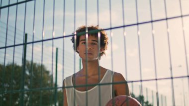 Basketball player training stadium zoom out. Curly hair young man playing ball at summer morning closeup. Stylish guy looking camera at empty playground portrait. Focused sportsman posing urban place 