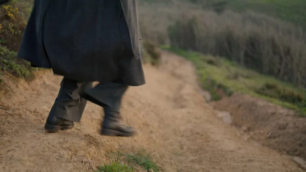 Woman legs running rural path in black casual boots close up. Unknown active dark-haired girl jogging on countryside road jumping. Back view energetic brunette wearing dark long coat. Activity concept