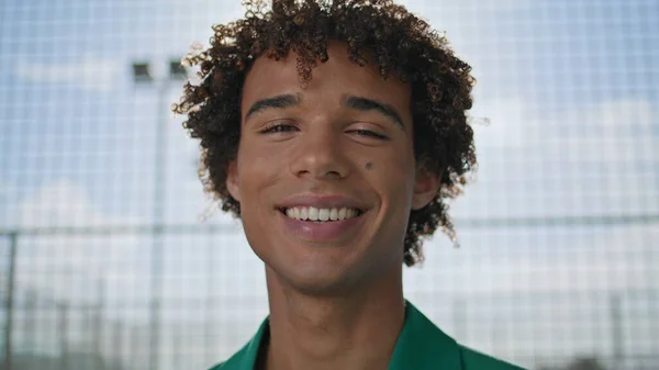 Positive teenager laughing street fashion portrait. Smiling young man standing looking camera alone closeup. Curly hair model demonstrating emotions at stadium. Cheerful guy face enjoying outdoors