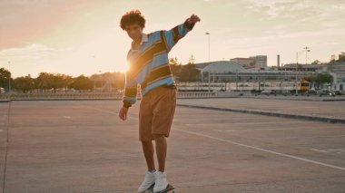 Active skateboarder making trick on board at city street. Curly hair sportsman rolling on asphalt alone. Young man foot stopping skate outdoor. Contemporary guy enjoying training at sunset place 