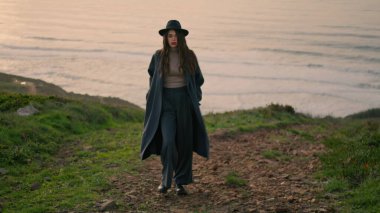 Energetic young lady walking coast in stylish coat with beautiful ocean backdrop. Attractive brunette in elegant hat posing at nature calm autumn evening. Confident woman enjoy cloudy landscape.