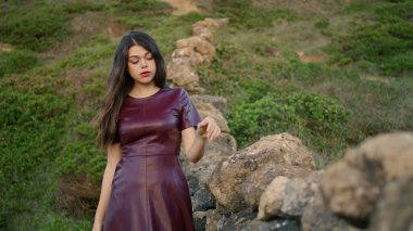 Gorgeous young woman relaxing walking hill green grass near rocky wall close up. Lonely romantic girl wearing dark dress going down slope looking camera. Gentle brunette enjoy calm leisure gloomy day.