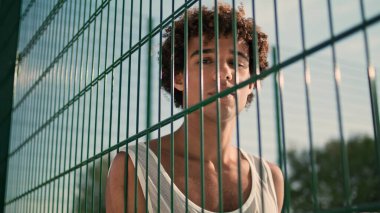 Young sportsman posing basketball court portrait. Curly bad guy looking camera with ball in hands alone. Serious teenager face staring lens on sport ground. Young man leaning stadium grid at summer 