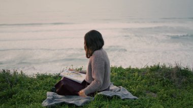 Young woman sitting seashore on blanket reading book at dramatic sea view. Attractive relaxed girl resting at beach in harmony with nature. Carefree brunette wearing sweater enjoy literature on shore.