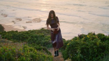 Dreamy romantic woman holding book walking green hill in front calm ocean evening time. Attractive long-haired girl wearing dark dress relaxing at beautiful seashore going on grass. Rest concept.
