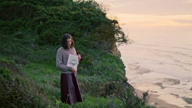 Beautiful brunette woman observing looking at wonderful overcast seashore nature. Young relaxed girl artist holding album with drawings cloudy evening. Calm lady spending weekend alone at gloomy beach