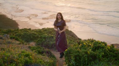 Calm brunette relaxing on seashore standing green grass hill wearing dark dress. Attractive long-haired dreamy woman holding book enjoying seascape evening time. Tranquil young girl posing on nature.
