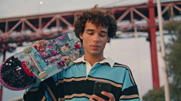 当代滑板手发短信手机户外特写 卷曲的年轻人带着长板在堤岸上的肖像 积极的家伙聊天智能手机日落桥的地方 英俊的少年步行城市 — 图库照片