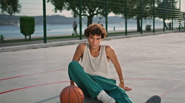 卷曲的运动员坐在体育馆里变大了 年轻的篮球运动员独自在露天休憩 一个自信的家伙在阳光灿烂的早晨看着相机 在街头肖像画前装腔作势的青少年 — 图库照片