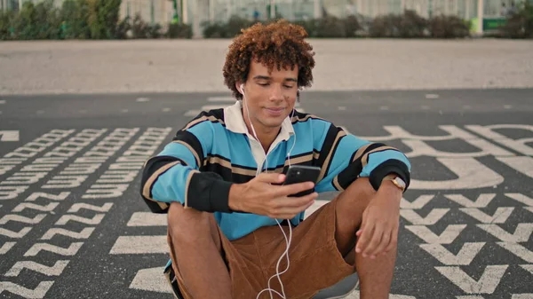 在街上的特写镜头里 笑着拍智能手机 耳机的家伙听音乐在手机室外放大 千禧年的嬉皮士在城市里表现得很酷 积极创造社交媒体内容的青少年 — 图库照片