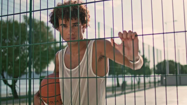 篮球运动员摆出体育场的肖像 卷发少年看着摄像头在城市的背景放大 当代学生紧紧抓住格子单独特写 穿着运动服的坏人享受夏天 — 图库照片