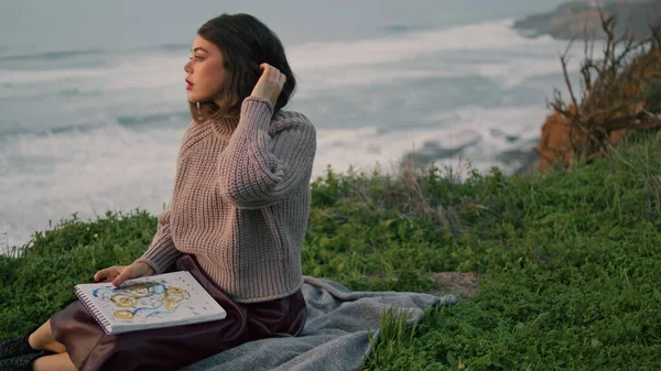 Calm dreamy woman relaxing on blanket with book looking evening ocean view. Attractive serious model posing sitting green grass seacoast at autumn dusk. Stylish thoughtful girl enjoy gray sea waves.