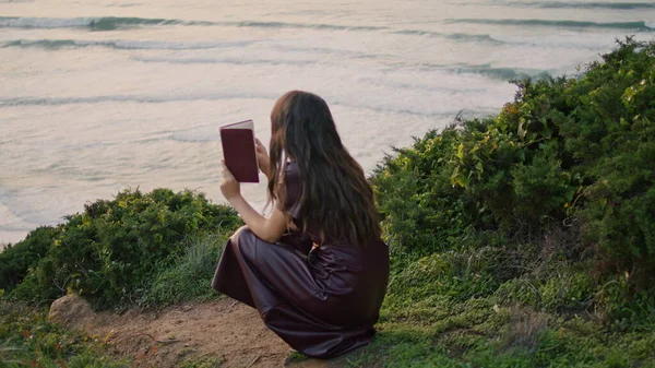Relaxed young woman holding book sitting in front beautiful ocean waves closeup. Attractive tranquil girl enjoy reading on nature cloudy autumn evening. Romantic brunette relax alone on green seashore