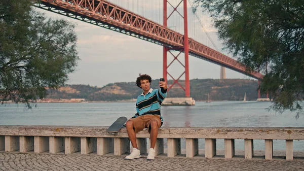 海岸のワイドショットでセルフィーを作るカーリースケーター 川の見える堤防でモバイルで座ってスタイリッシュな観光客 リスボンポルトガルでスマートフォンを使う現代人 有名な場所で千年ブロガー — ストック写真