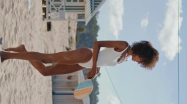 Plajdaki kız sahilde raket tutuyor. Sıska seksi bikinili kadın, hafta sonu bilinmeyen partneriyle dikey olarak eğleniyor. Afro-amerikalı oyuncu seyahat aktivitelerini seviyor