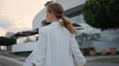 Kaygısız kız akşam vakti sokakta yürüyor. Mutlu, gülümseyen bir kadın zarif beyaz takım elbisesiyle modern kentsel alanda geziniyor ve mutluluğu hissediyor. Neşeli genç bayan hafta sonunun tadını çıkarın.