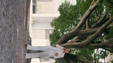 Kendine güvenen bayan patron kaldırım kenarında yürüyor ve güneşli bir günde telefonla dikey çekim yapıyor. Harika bir iş kadını işe gitmek için acele ediyor. Zarif bir genç kadın telefonun altında ağaçların arasında geziniyor.