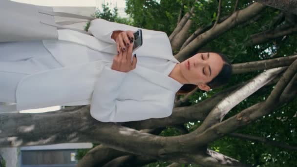 美しい緑の木の下を垂直に歩いているビジネスマンダイヤル電話番号 ホワイトスーツのエレガントなシックなビジネスレディー電話 スマートフォンワークコミュニケーションを使用して真剣な豪華な女性 — ストック動画