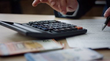Unknown man using calculator counting corporate budget indoors close up. Male hands pressing calculator buttons analyzing business income. Accountant working at table with documents bills money cash.