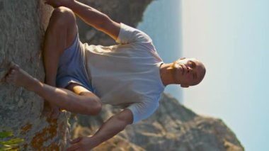 Sabah güneşinde kaslı bir adam Asana egzersizi yapıyor. Okyanus yamacında gerinen bir sporcu. Yoga pozisyonunda nefes alan güçlü kişi dikey görünüm