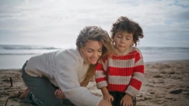 Anne çocuk kumsal kapanışı oynuyor. Gülümseyen bir ebeveyn okyanus kıyısında oğlunu öperken eğleniyor. Sevecen anne anaokulu çocuğuyla aşk dolu bir hafta sonu geçirdiğini ifade ediyor. Aile bağları.