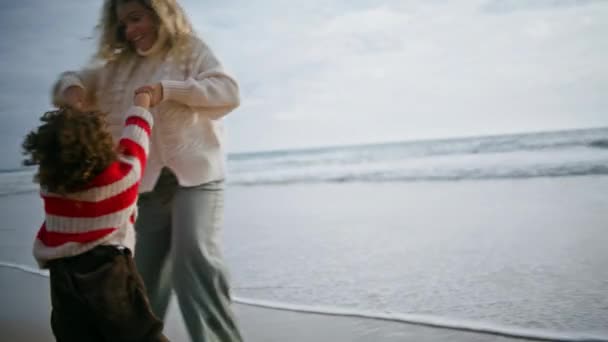 快乐的母亲在海滨纺纱儿子 快乐的家庭在秋天的假期里玩着海滩放松 漂亮的微笑保姆在旋转与小卷曲男孩玩乐 在一起的快乐时光 — 图库视频影像