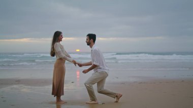 Amorous lovers engaging at sunset seashore. Handsome man making surprise marriage proposal to beloved woman at evening beach. Rejoicing girl saying yes. Emotional moment. Relationship goals concept