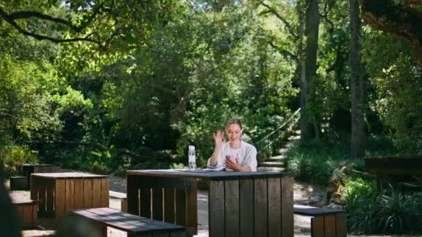 緑の公園の木のピクニックテーブルに座っているスマートフォンのカメラで挨拶する美しい女性 晴れた森でリラックスする魅力的な少女ビデオ 携帯電話でBogを記録するヘッドフォンで幸せな女性 — ストック動画