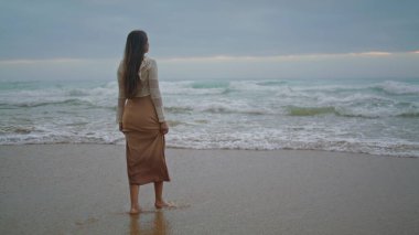 Romantic girl looking ocean view at evening. Dreamy woman walking on stormy beach wide shot. Female model spending vacation at seaside alone. Brunette lady enjoying of scenery marine landscape