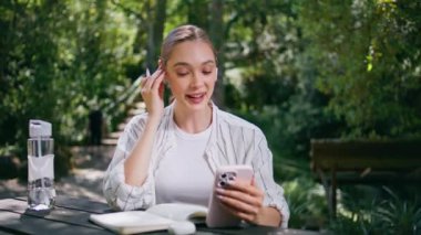Kulaklıklı bir öğrenci akıllı telefondan Webinar dinliyor Green Park Doğa 'ya yakın oturuyor. Kulaklıkla bilgi dinleyen mutlu bir kadın not defterine not alıyor. Bahçeden güzel kız videosu arıyor..