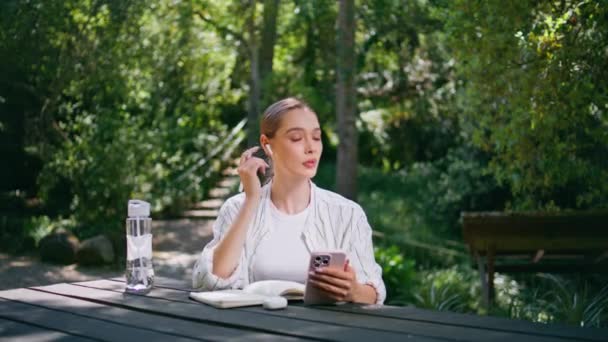 戴着耳机的快乐女人坐在绿色公园的桌子旁边接着视频电话 快乐的女孩对着手机摄像机打招呼 在阳光普照的自然中放松 可爱的女士在森林里遥谈 — 图库视频影像