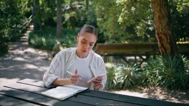 木のテーブルの森に座っているスマートフォンからコピーブックに情報を指摘する少女作家 携帯電話のインターネットを使ってエッセイを作成する熟練した学生 緑の自然の中でモバイルアプリを勉強している女性 — ストック動画