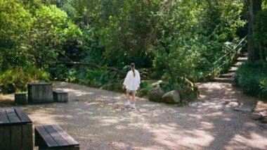 Orman dinlenme yerinde yürüyen bir kadın taze yemyeşil yeşilliklere bakıyor. Tasasız genç bayan yeşil güneşli parkta tek başına yaz haftasonunun tadını çıkarıyor. Mutlu hayalperest kız doğada dinleniyor, özgürlüğü hissediyor..