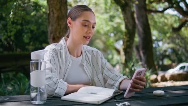 女自由职业者通过智能手机远程咨询客户 坐落于绿林景观中 快乐的微笑女孩视频电话通过手机复制关于阳光自然的书 女士在网上聊天 — 图库视频影像