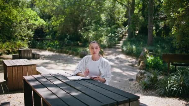 ノートパッド付きの木製テーブルに座っている森で働く魅力的なフリーランサー 自然の中でスマートフォンを使用してオンラインで情報を探しているリラックスした若い女性 研究のためのインターネットサーフィンのリラックスペンギン学生 — ストック動画
