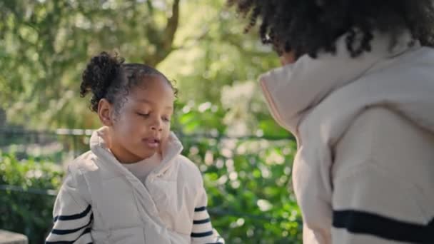 小女孩和可爱的妈妈一起坐在长椅绿色的花园里 漂亮的小可爱享受春天里阳光灿烂的家庭周末 非洲裔美国人女儿和不认识的妈妈在一起放松一下 — 图库视频影像