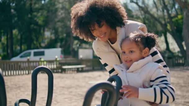 アフリカ系アメリカ人の子供がカルーセルで遊んでおり 母親が近づいている 笑顔の母親は 遊び場でメリーゴーラウンドから女の子を取ります 公園で娘と楽しく過ごしている幸せな女性 アクティブな子供時代のコンセプト — ストック動画