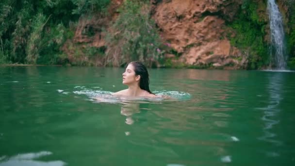 美しい夏の自然の中で裸のモデルスイミングクリフ湖 パラダイスバケーションを一人で楽しむカメラを見ているキャリーフリーの女性 トロピカルラグーン ウォーターベイで休むスリムな少女 小さな滝の背景 — ストック動画