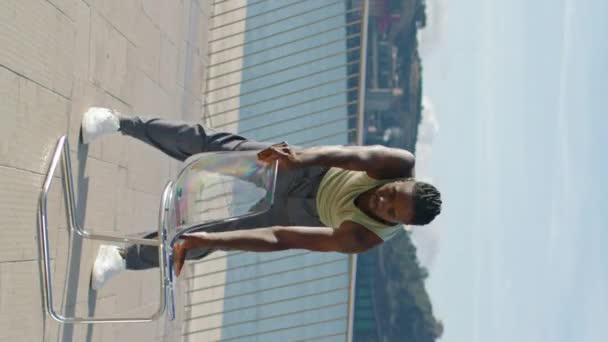柔軟なドレッドパフォーマーダンスチェアエンバンクメント垂直ビデオ バレエのテクニックを示すバーレスクダンサー アフリカ系アメリカ人の優雅な男性が振付海の景色を演じています プロの都市ダンス — ストック動画